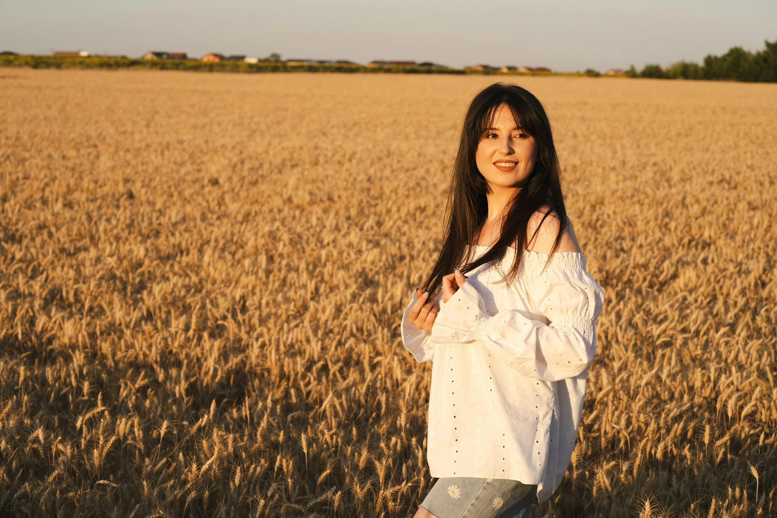 A young woman with long dark hair stands in a golden wheat field during sunset, wearing a white off-the-shoulder blouse and gray shorts with flower embroidery, smiling and looking slightly to the side.