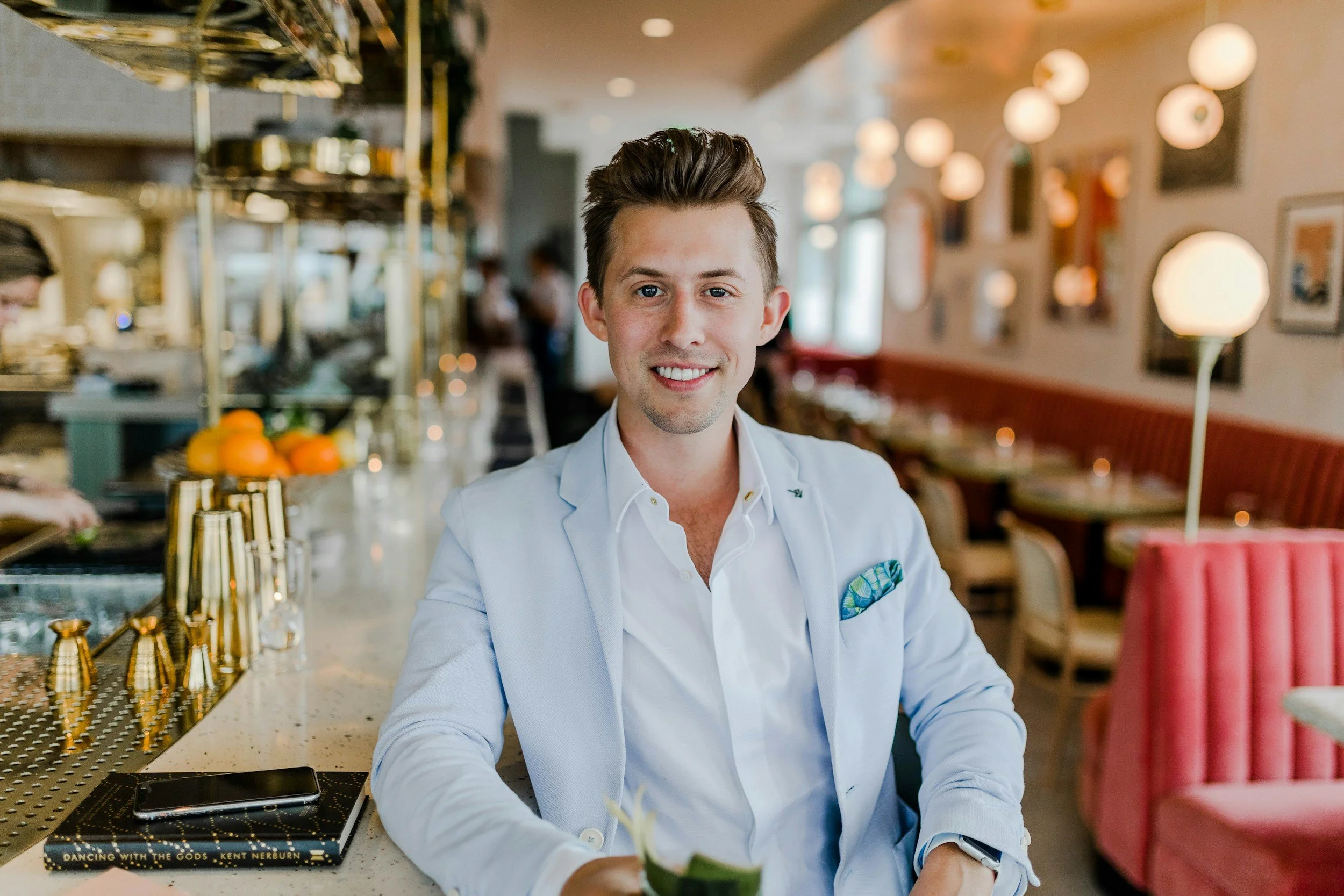 A smiling young man in a light blue blazer and white shirt sitting at a restaurant bar or counter with a decorative and colorful interior in the background.
