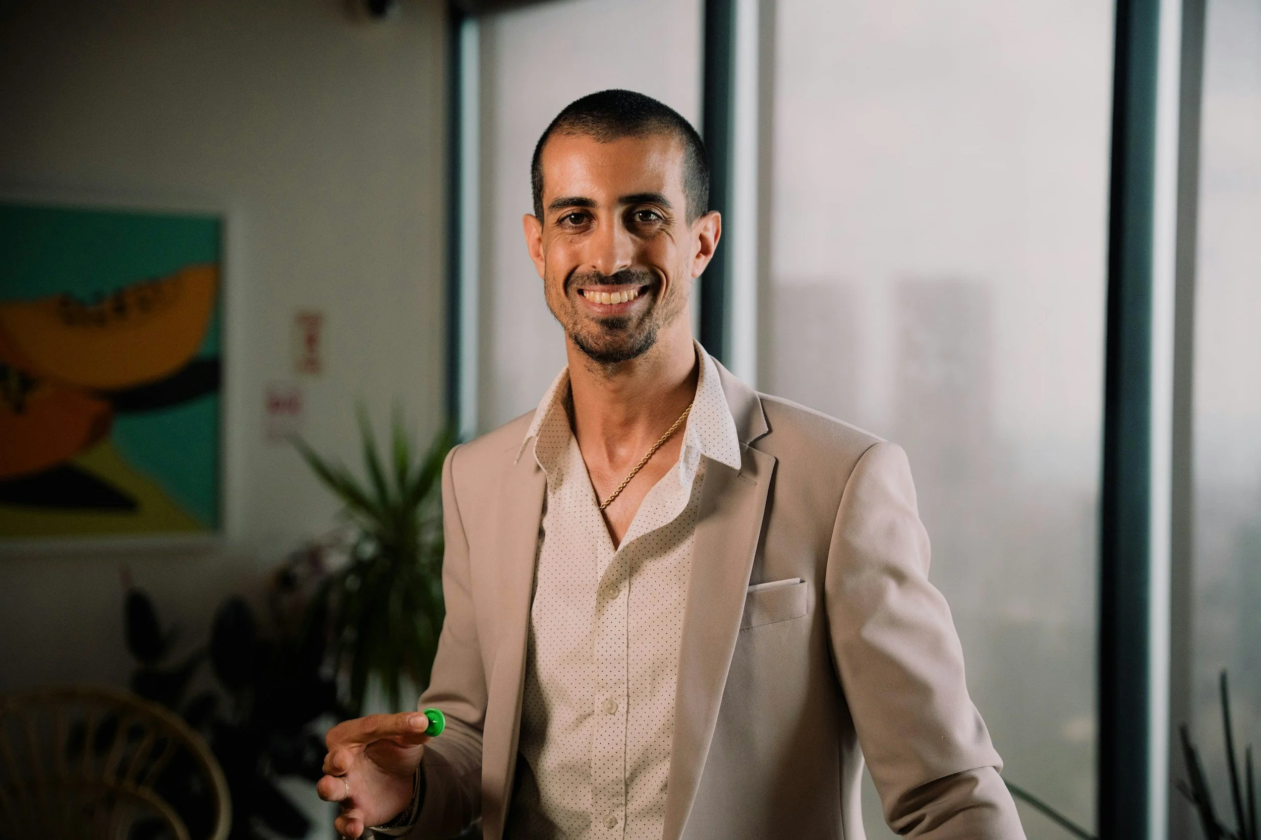 A smiling man with short dark hair and a beard, wearing a light beige suit, white shirt, and gold chain. He is holding a green pen and standing indoors with large windows and artwork in the background.