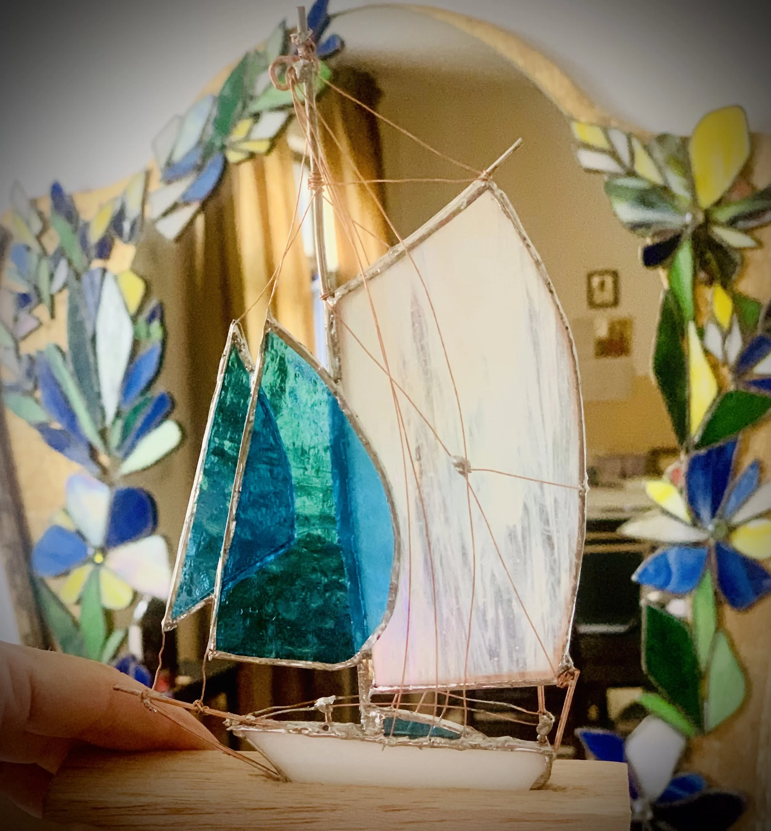 A stained glass sailboat with blue and white sails, surrounded by colorful flowers and sailboat frames, displayed indoors.