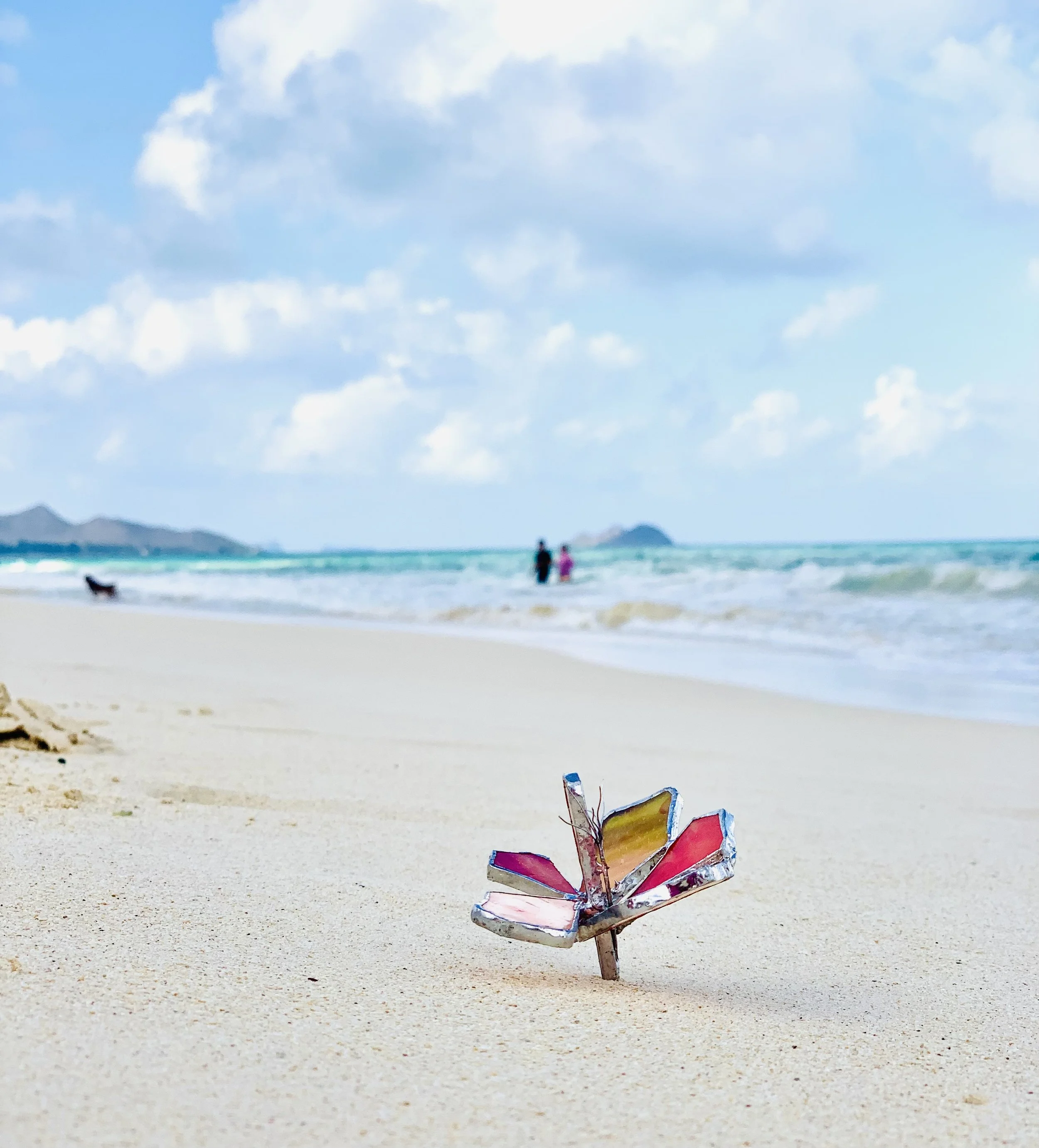 Hawaiian flower made of colored glass in Tiffany technique, on Waimanalo Beach from Oahu, with white sand, calm, turquoise waters, and beautiful backdrop.