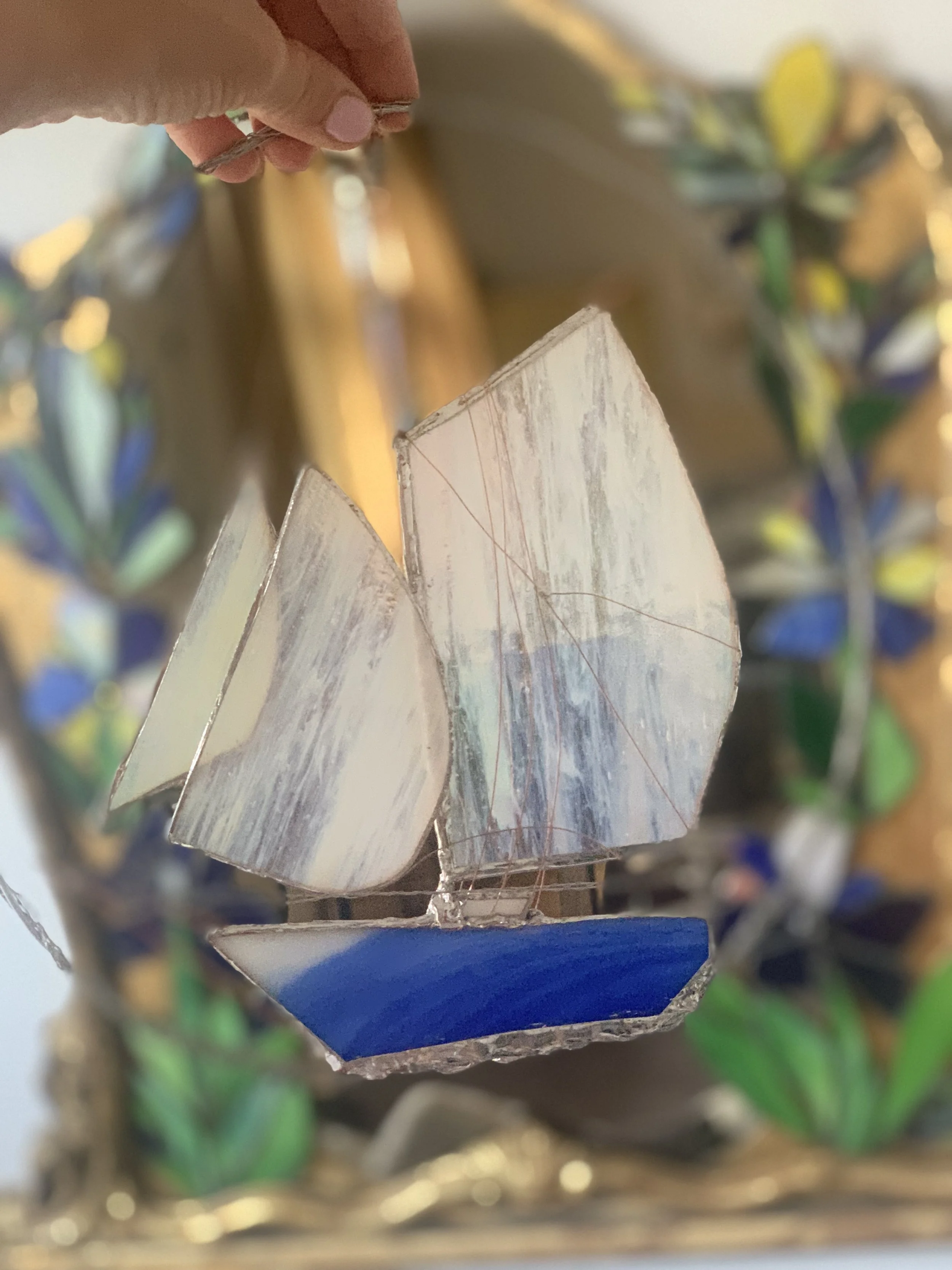 Close-up of stained glass art depicting a sailboat with white and blue glass, with a background of a decorative structure featuring colorful stained glass pieces and gold accents.