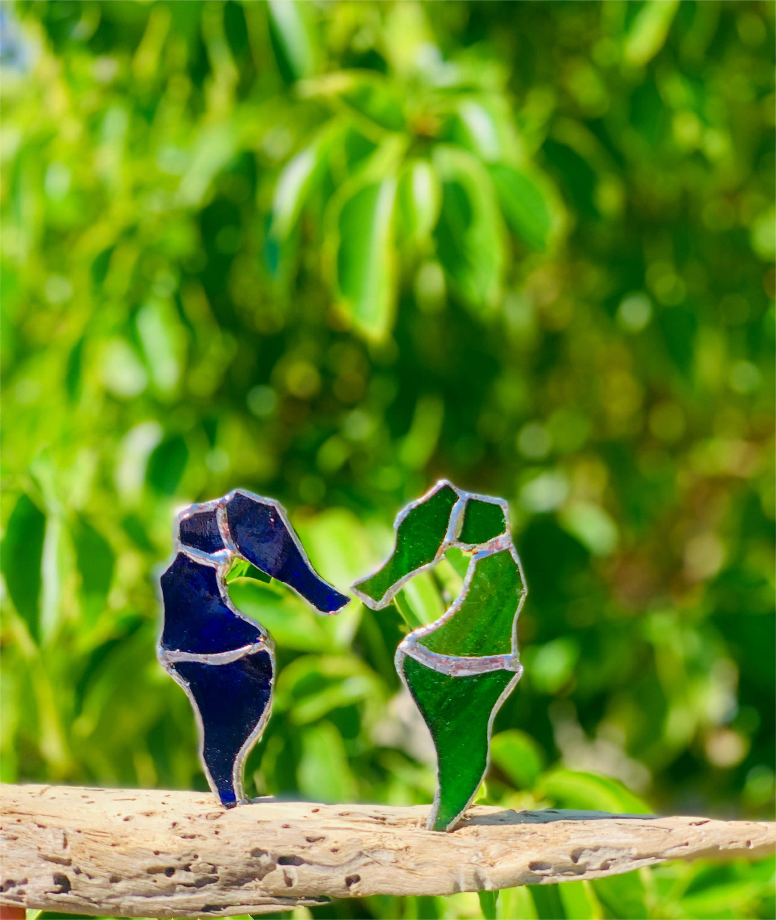 Colorful stained glass seahorses on a wooden branch with green foliage in the background.