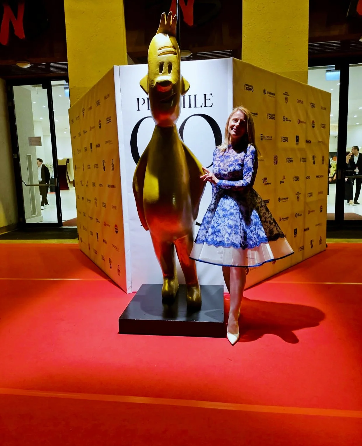 Artist Marie A radiates elegance in a blue lace dress with a petticoat, posing gracefully beside the iconic gold GOPO statue at a prestigious red-carpet event. A refined moment blending art, style, and cinematic glamour.