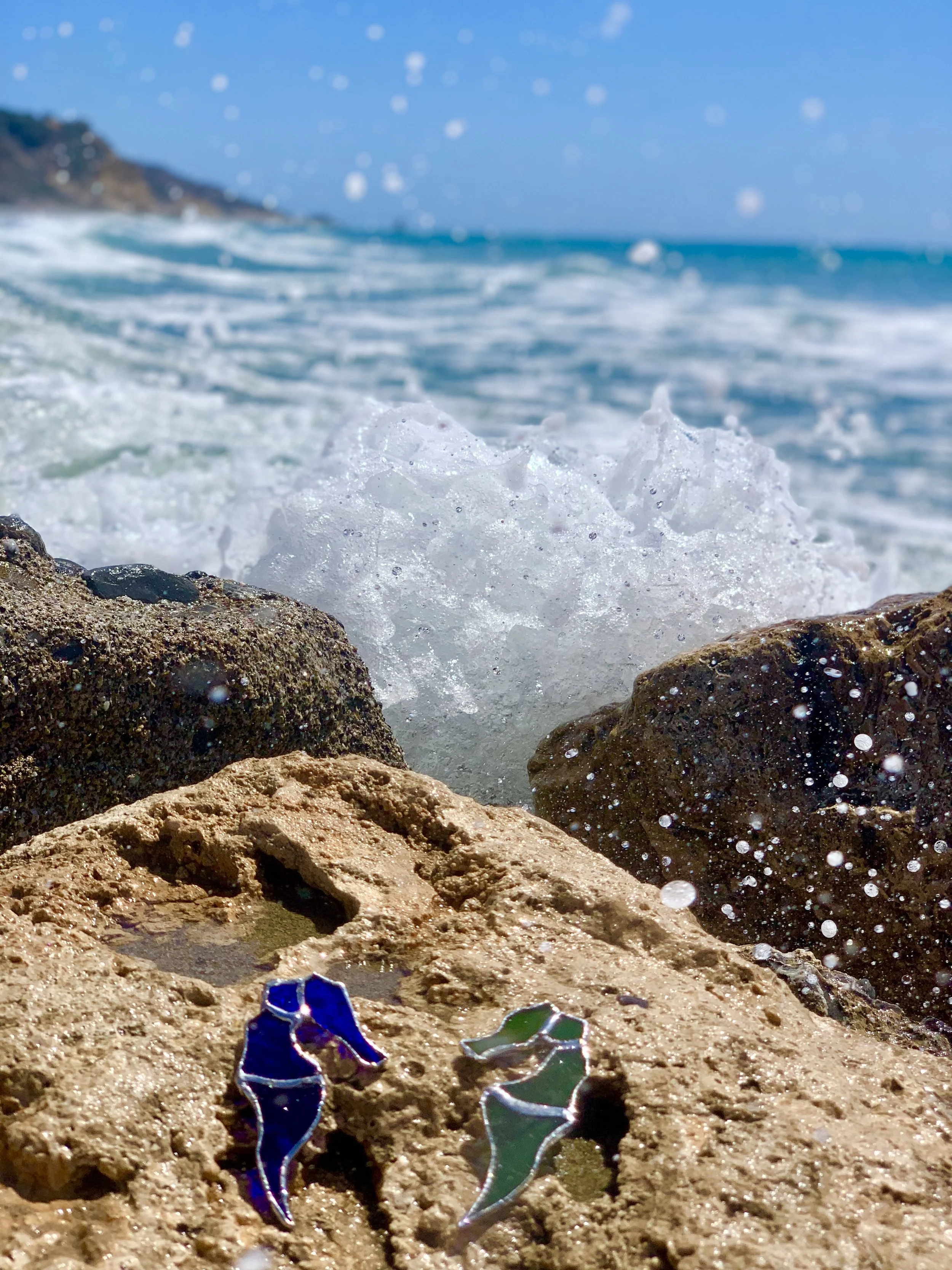 Close-up of textured brownish rocks on a beach with two small, colorful, glass ornaments in the foreground representing two sea horses. Ocean waves crashing in the background and a blue sky with scattered clouds.