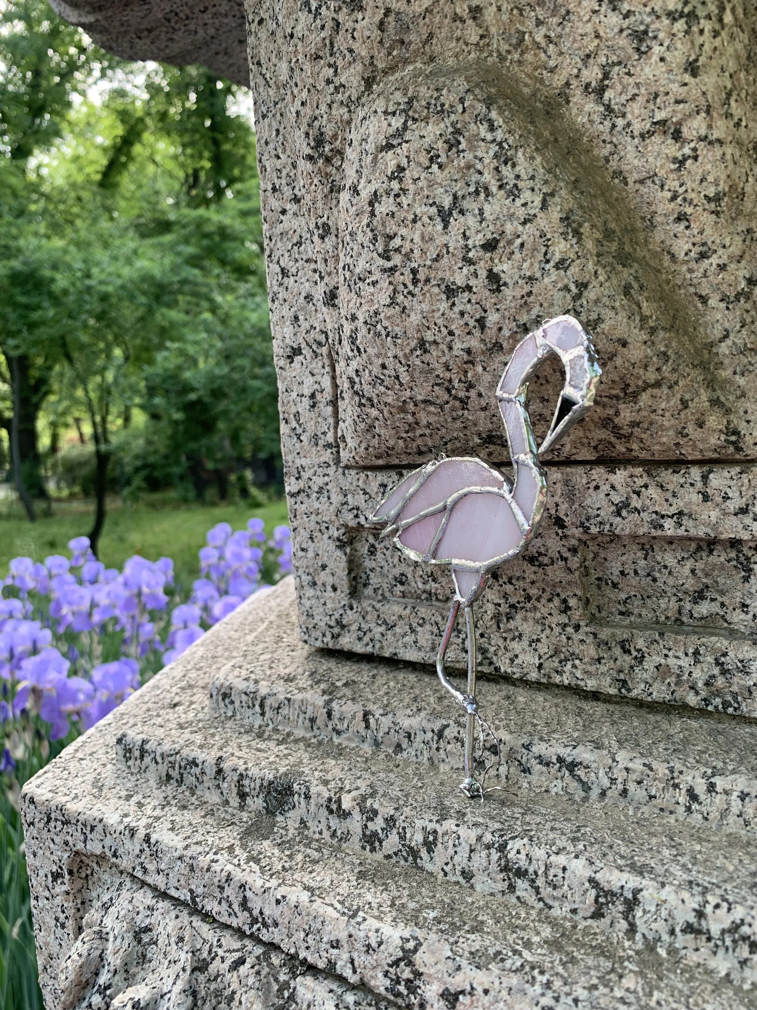 A stained glass ornament of a flamingo stained glass, near a stone monument, with purple flowers and green trees in the background.