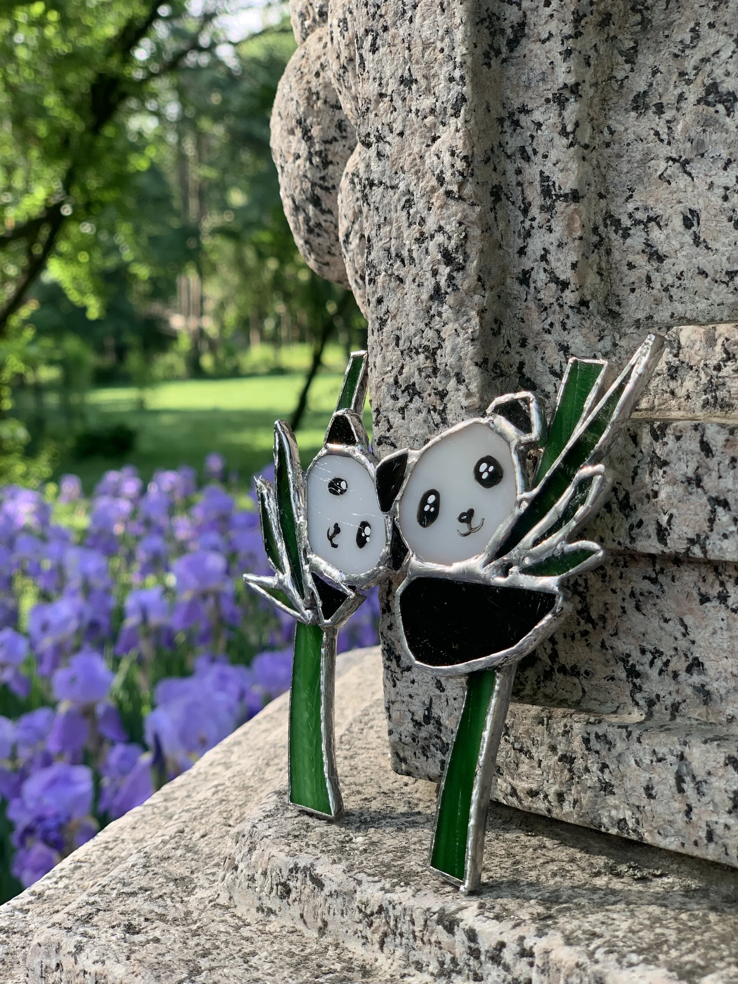 Colorful panda-shaped stained glass decoration attached to a stone structure, with a background of purple flowers and greenery.