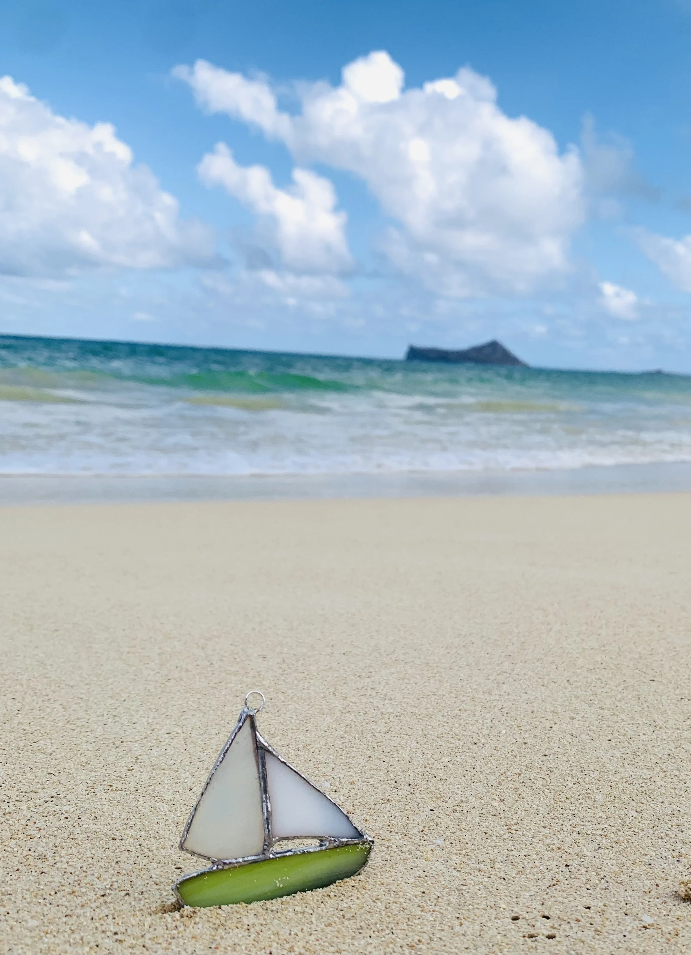 A small decorative sailboat made of stained glass on a sandy beach with ocean waves and a distant island under a partly cloudy blue sky.
