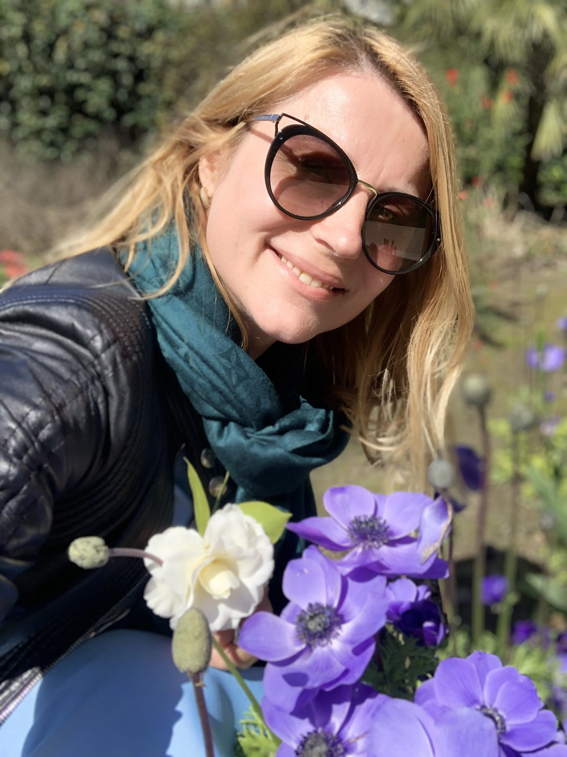 Artist Marie A radiates elegance, smiling while taking a selfie among purple and white flowers in a garden.