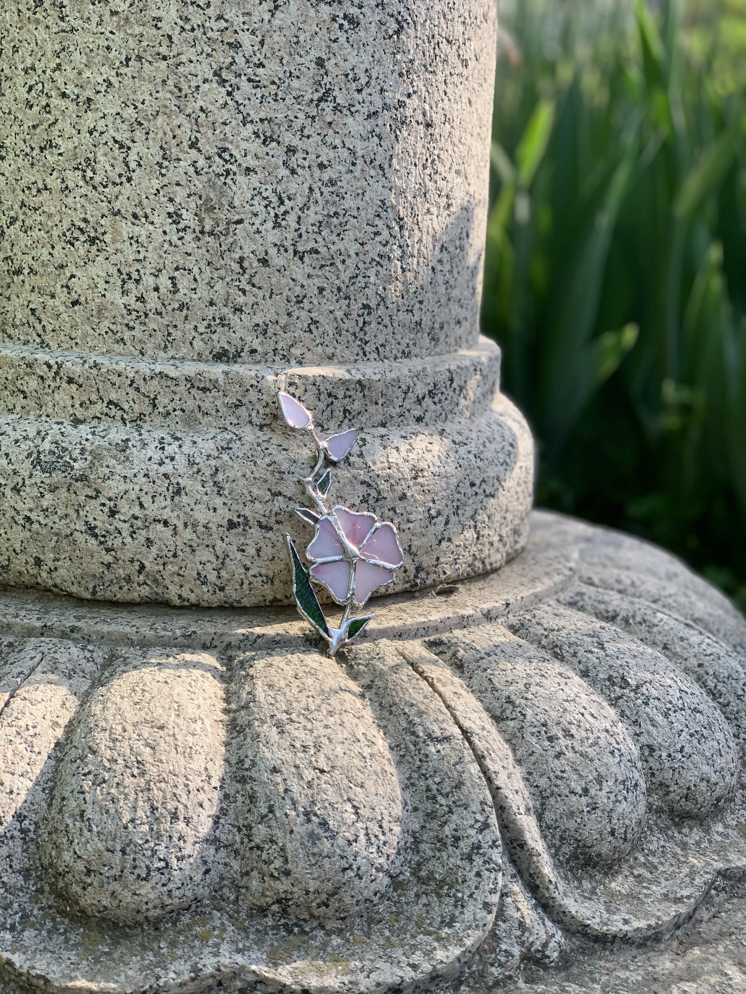 Tiffany technique necklace made of colored glass, with a pink and green floral pendant, set on an outdoor stone sculpture with green plants in the background.