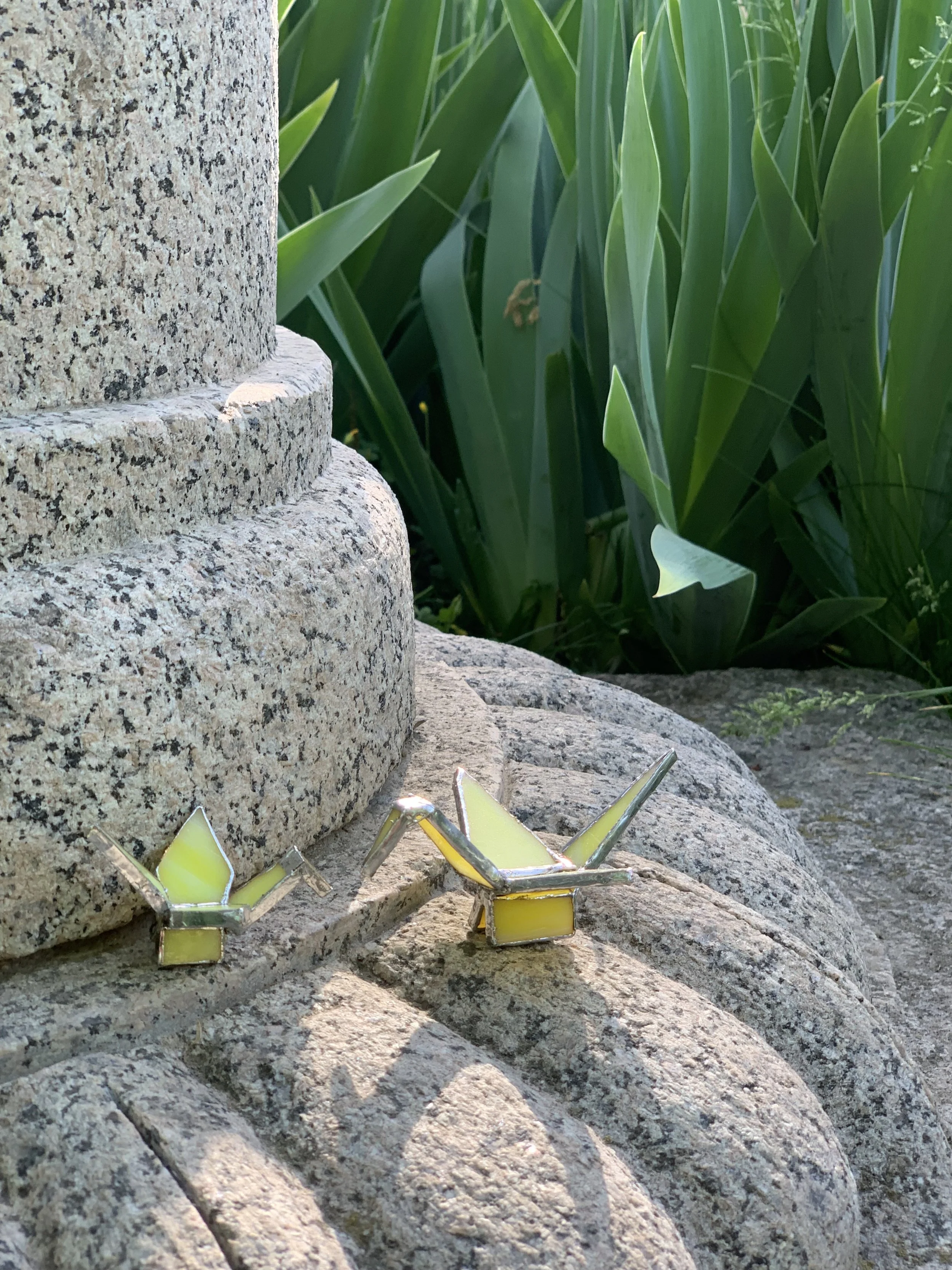 Two origami figurines representing two colored glass cranes, in the Tiffany technique, on a stone surface next to green plants in an outdoor setting.