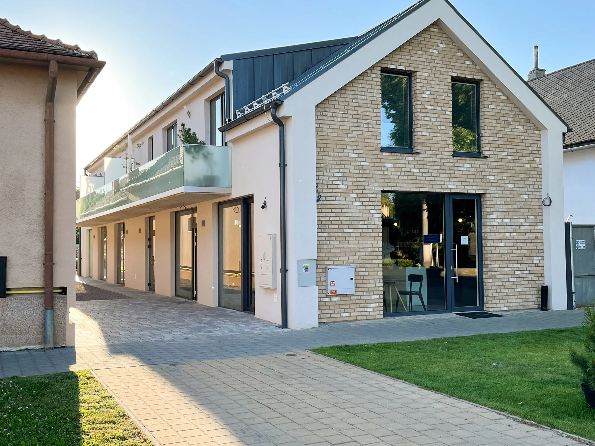 Modern residential building with brick facade, glass balcony, and large windows, set in a landscaped area with a pathway and lawn.