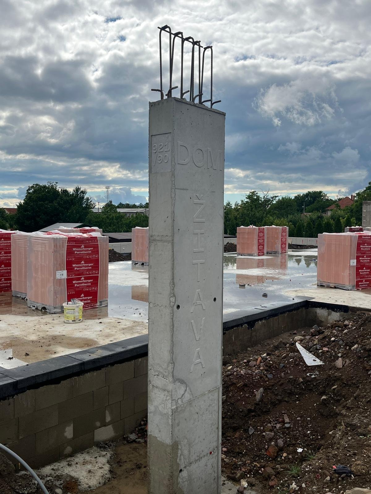 Concrete construction site with exposed rebar, marked pillar, and packaged building materials in the background under cloudy sky.