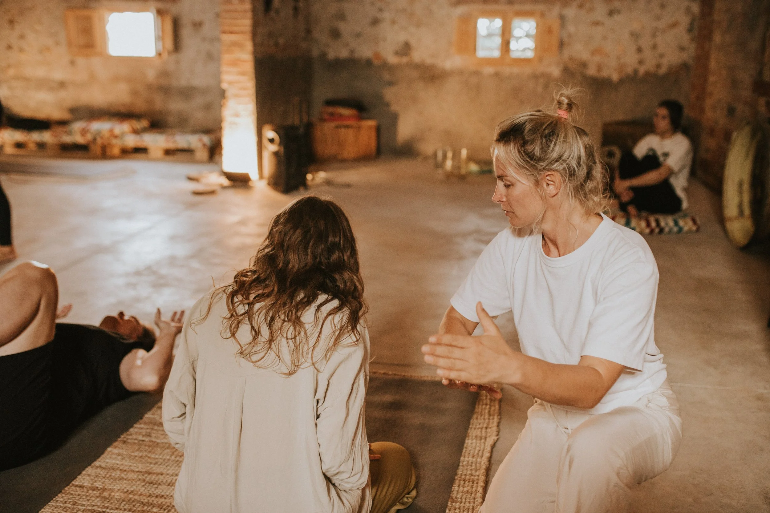 People engaged in a meditation or wellness session in a rustic room, with one person lying on the floor and others seated.