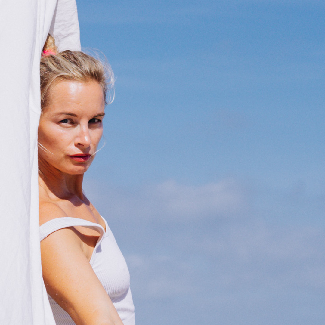 Woman in white top standing against a blue sky backdrop, partially obscured by a white curtain.