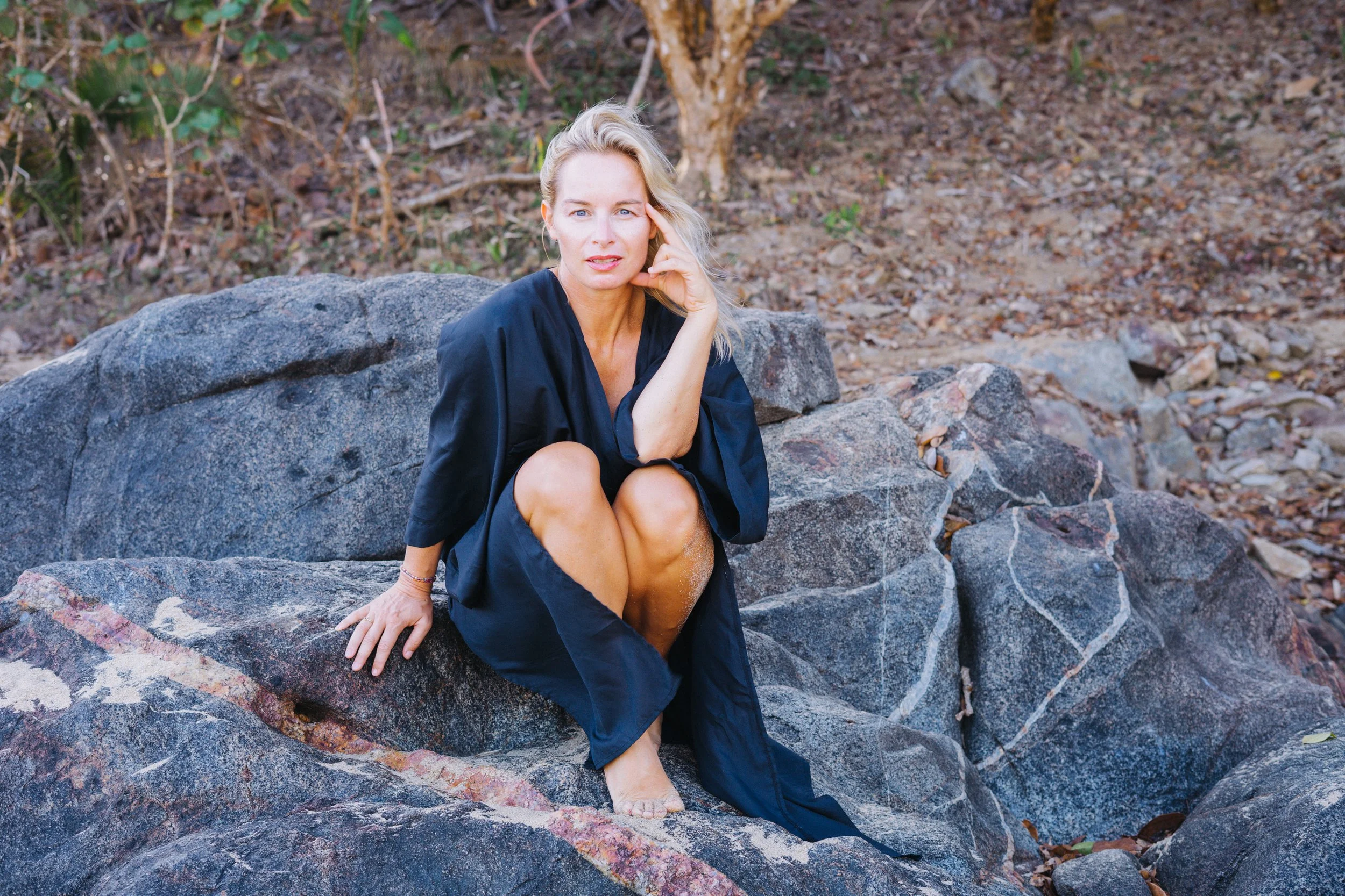 A woman in a navy blue dress sitting on large rocks in an outdoor setting, surrounded by dry foliage and bare trees.