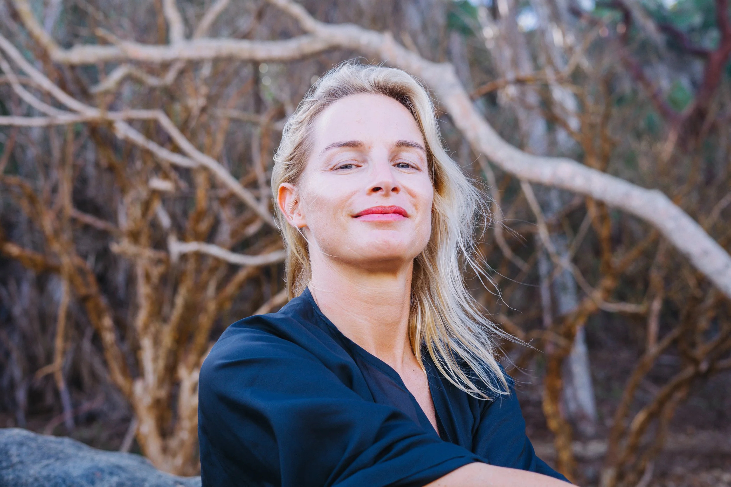 Eine Frau mit blonden Haaren in einem schwarzen Oberteil steht vor einem Hintergrund aus verworrenen Ästen im Freien.