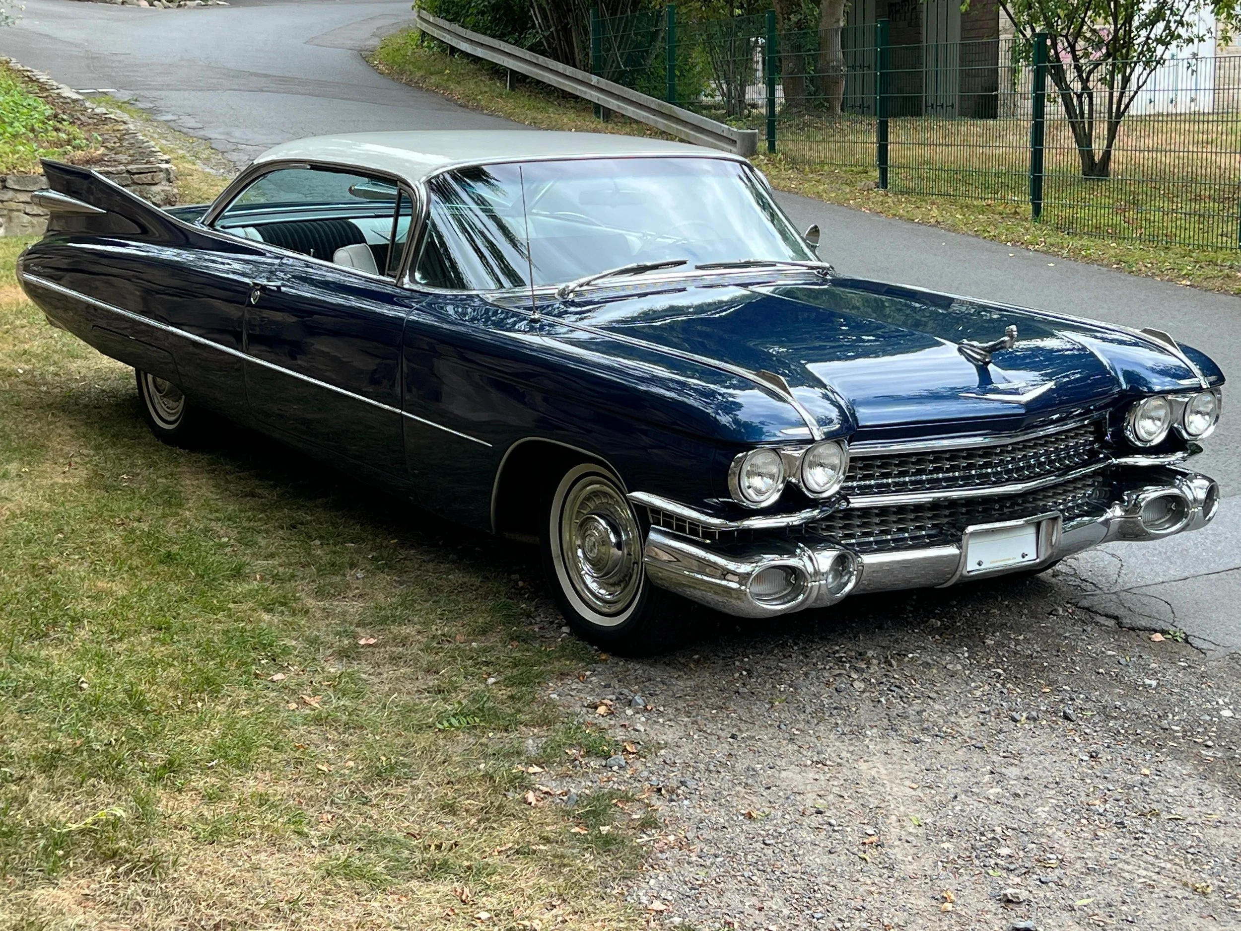 Ein klassischer dunkelblauer Cadillac Oldtimer mit weißem Dach, geparkt am Straßenrand auf grasigem Untergrund, mit Straßen und Bäumen im Hintergrund.