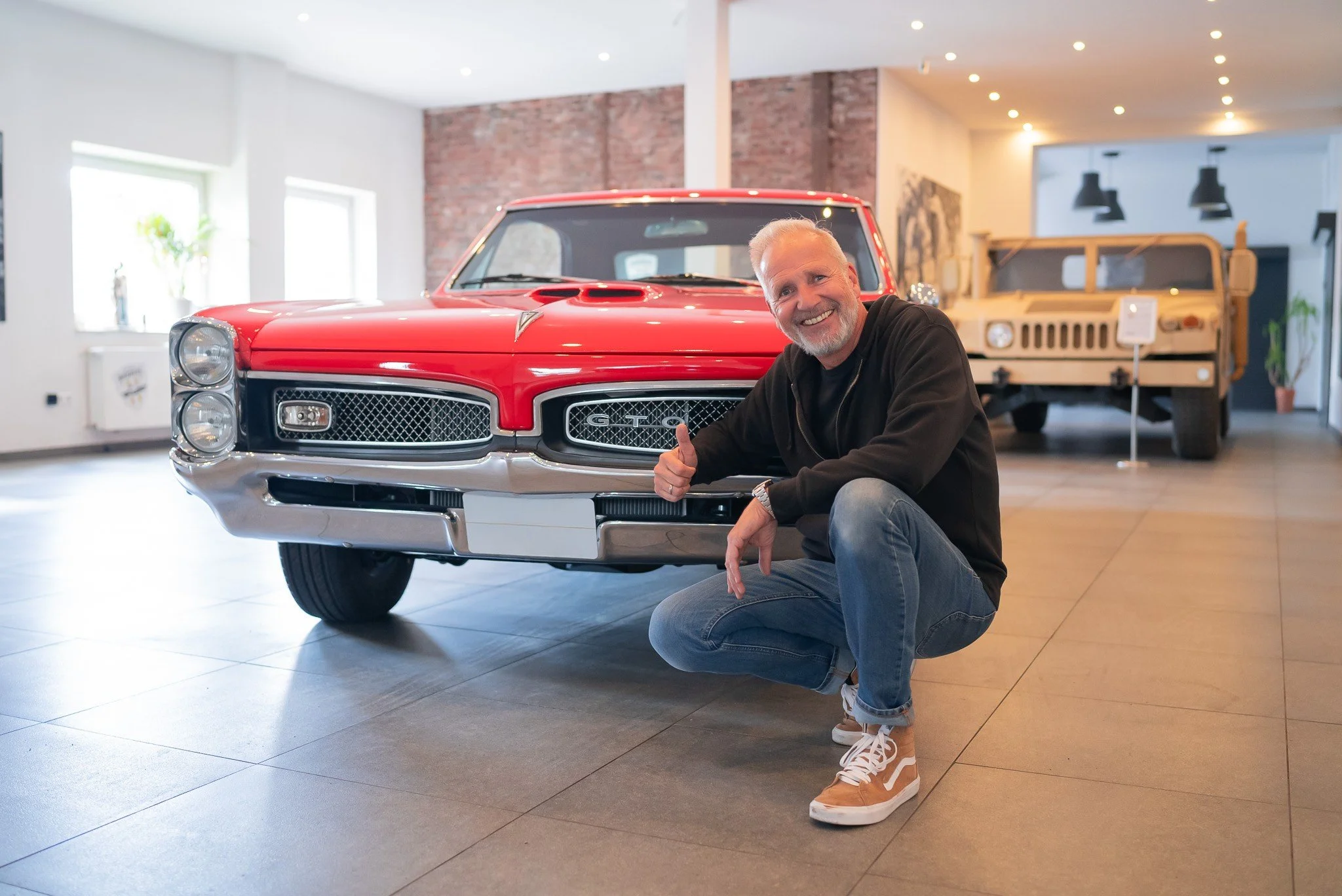 Peter Dornheim im Black-Hoodie, blauen Jeans und braunen Sneakers kniet vor einem roten Pontiac GTO!