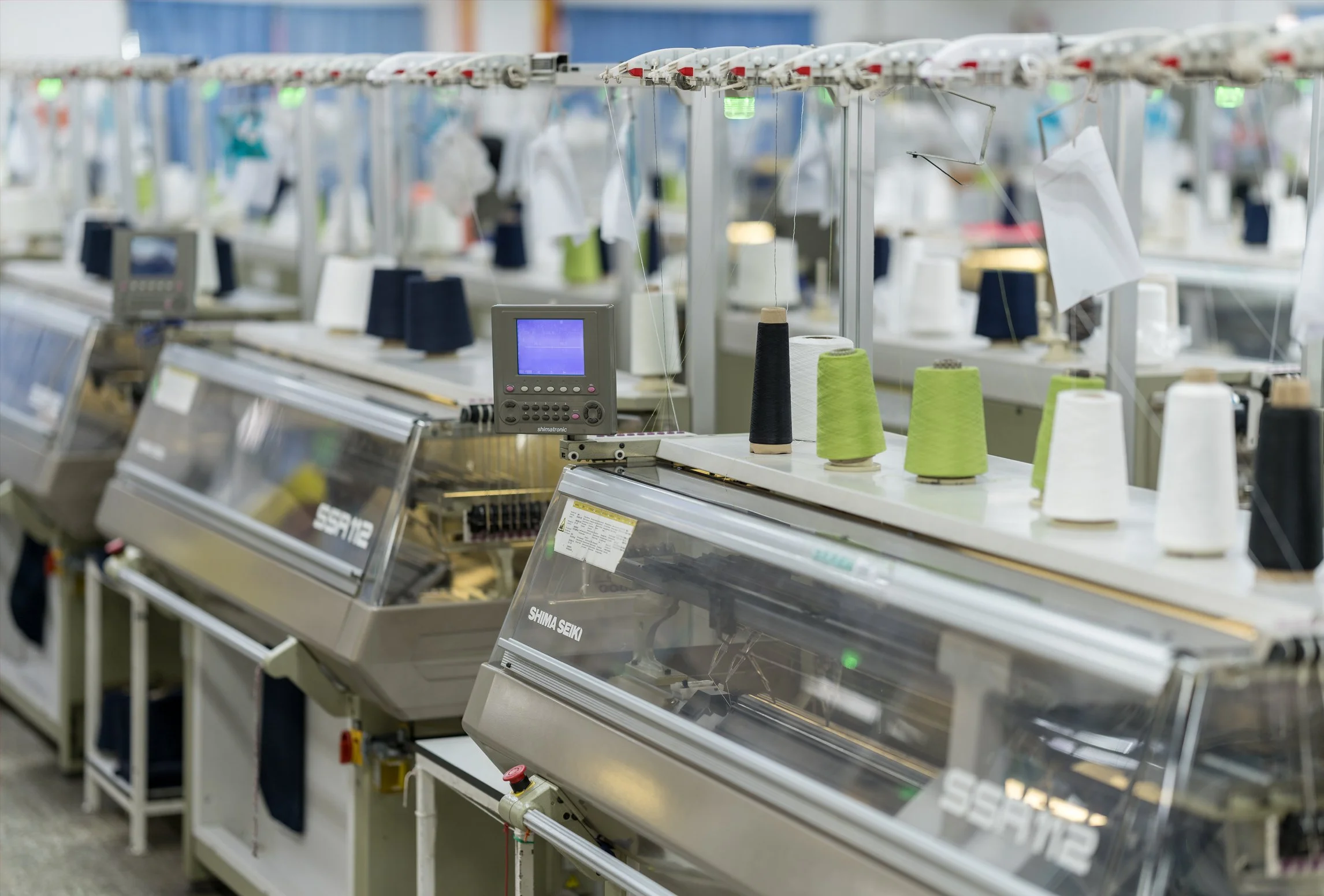 Textile factory with multiple sewing machines, cones of thread in white, black, and green, and digital screens for control.