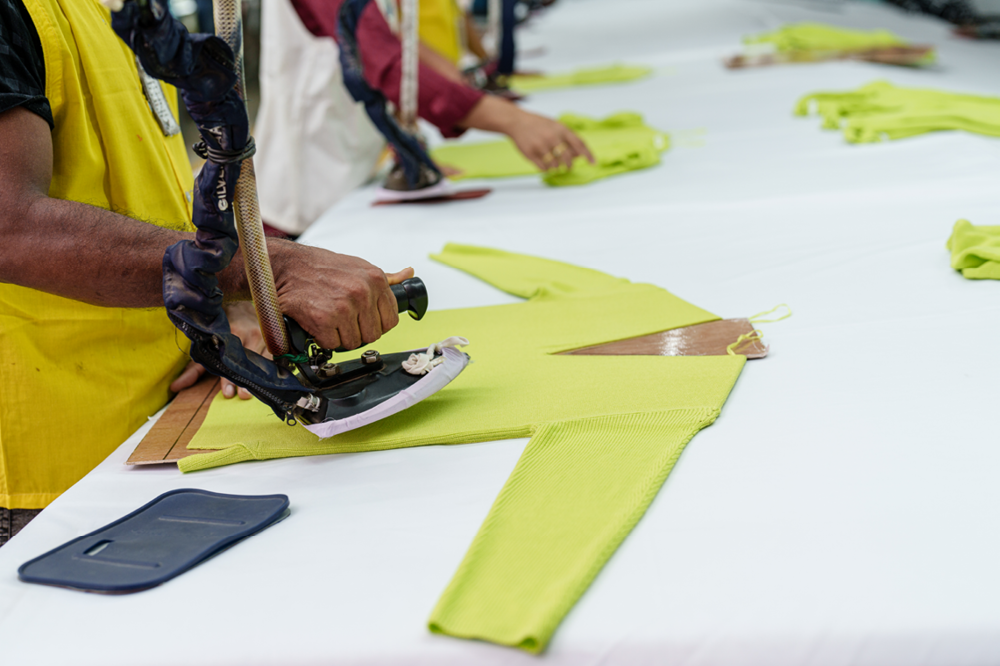 Person using an iron to press bright yellow fabric on a long table with other people working on similar projects.