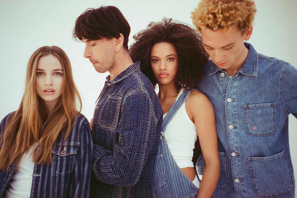 Four young adults standing closely, looking serious, against a plain background.