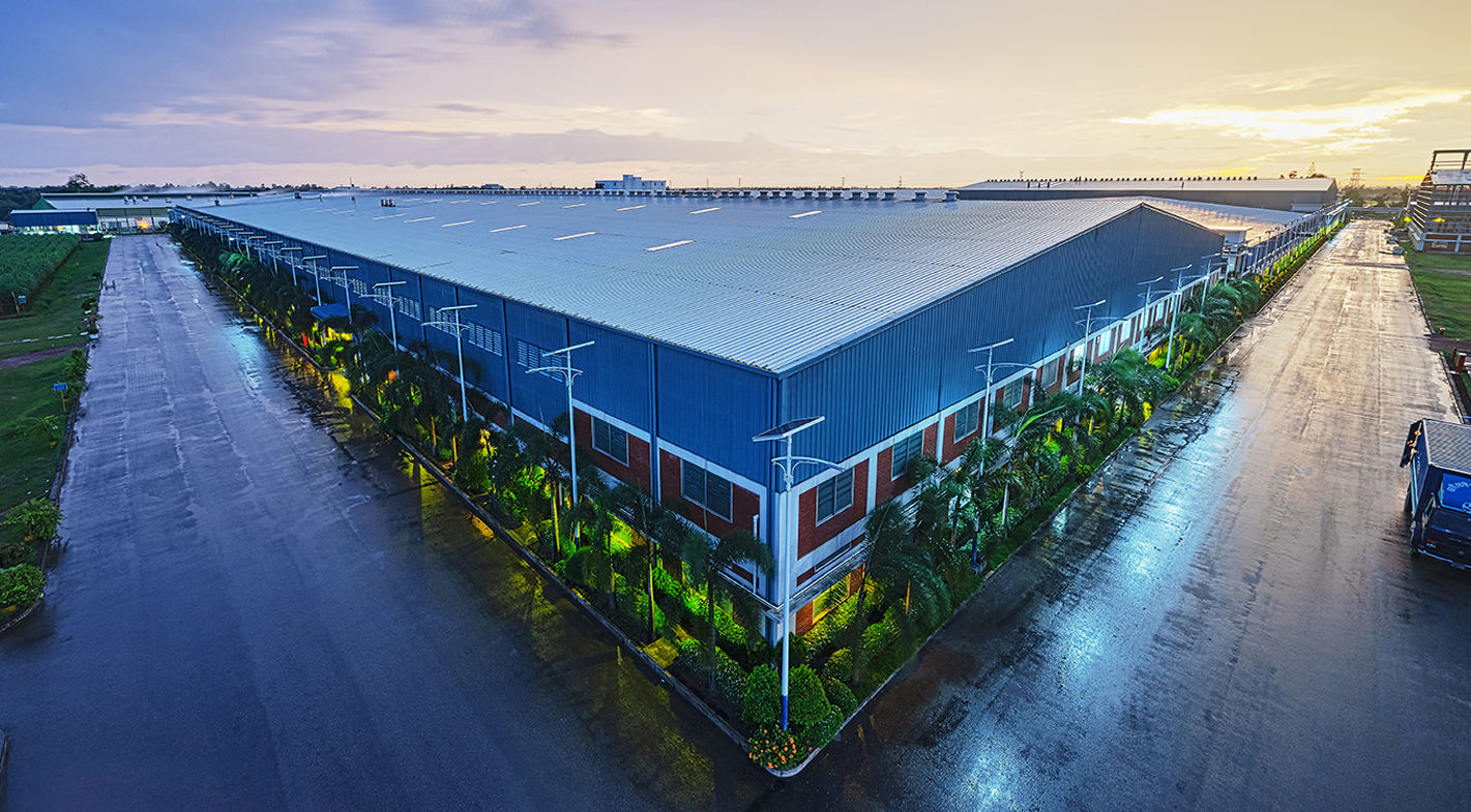 A large industrial warehouse with a metal roof, surrounded by streetlights, palm trees, and wet pavement at sunset.