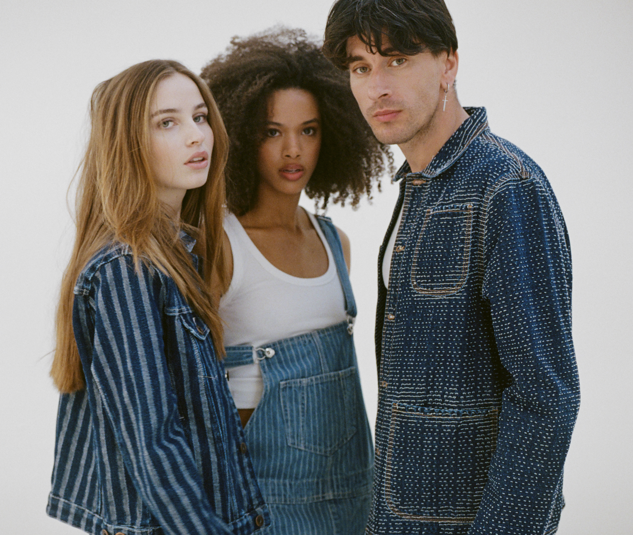 Three young adults, two women and one man, standing close together against a plain, light-colored background. They are wearing denim outfits and look directly at the camera.