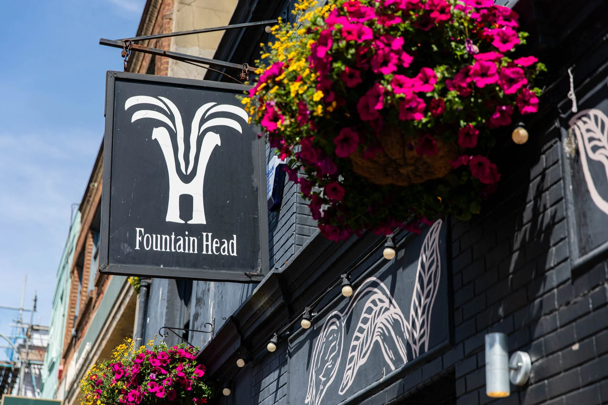 A black sign with white art of a fountain and the words "Fountain Head" hanging outside a building with pink and yellow flowers in the flower box above.