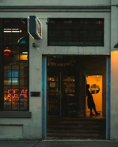A person wearing a hat and skirt standing inside a store, viewed through an open doorway, with warm yellow light illuminating the interior, contrasted by the cool exterior of the building.