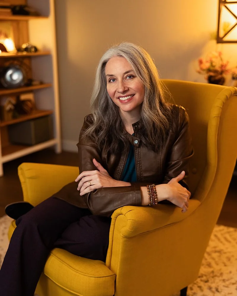 A woman with long gray hair sitting in a yellow armchair, smiling at the camera, in a warmly lit room with shelves and decor in the background.