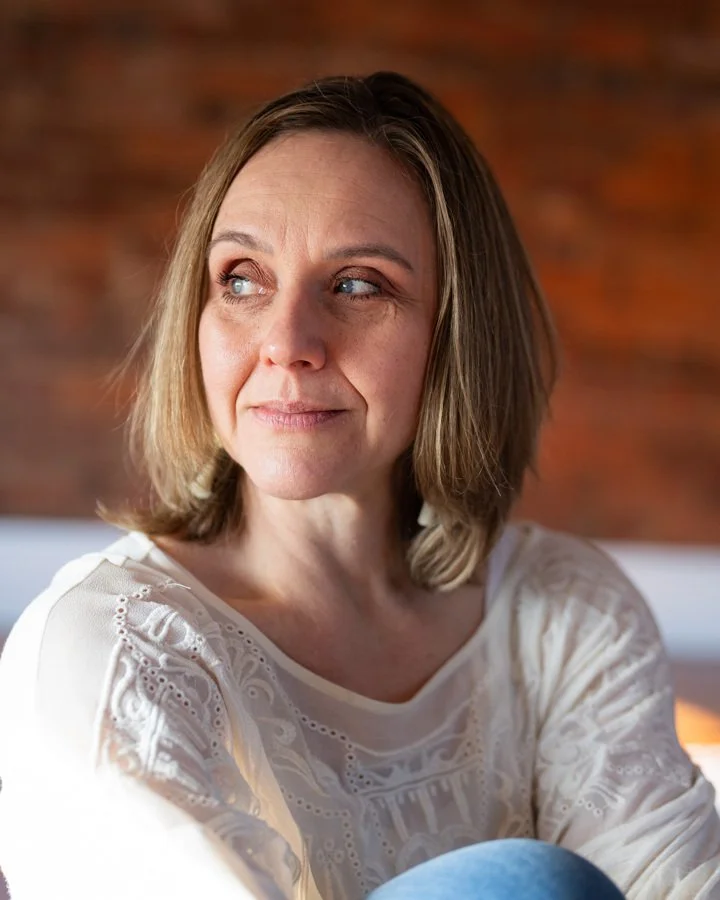Beautiful woman with short brown hair looking to the side, wearing a white patterned blouse, sitting indoors with a brick wall background.