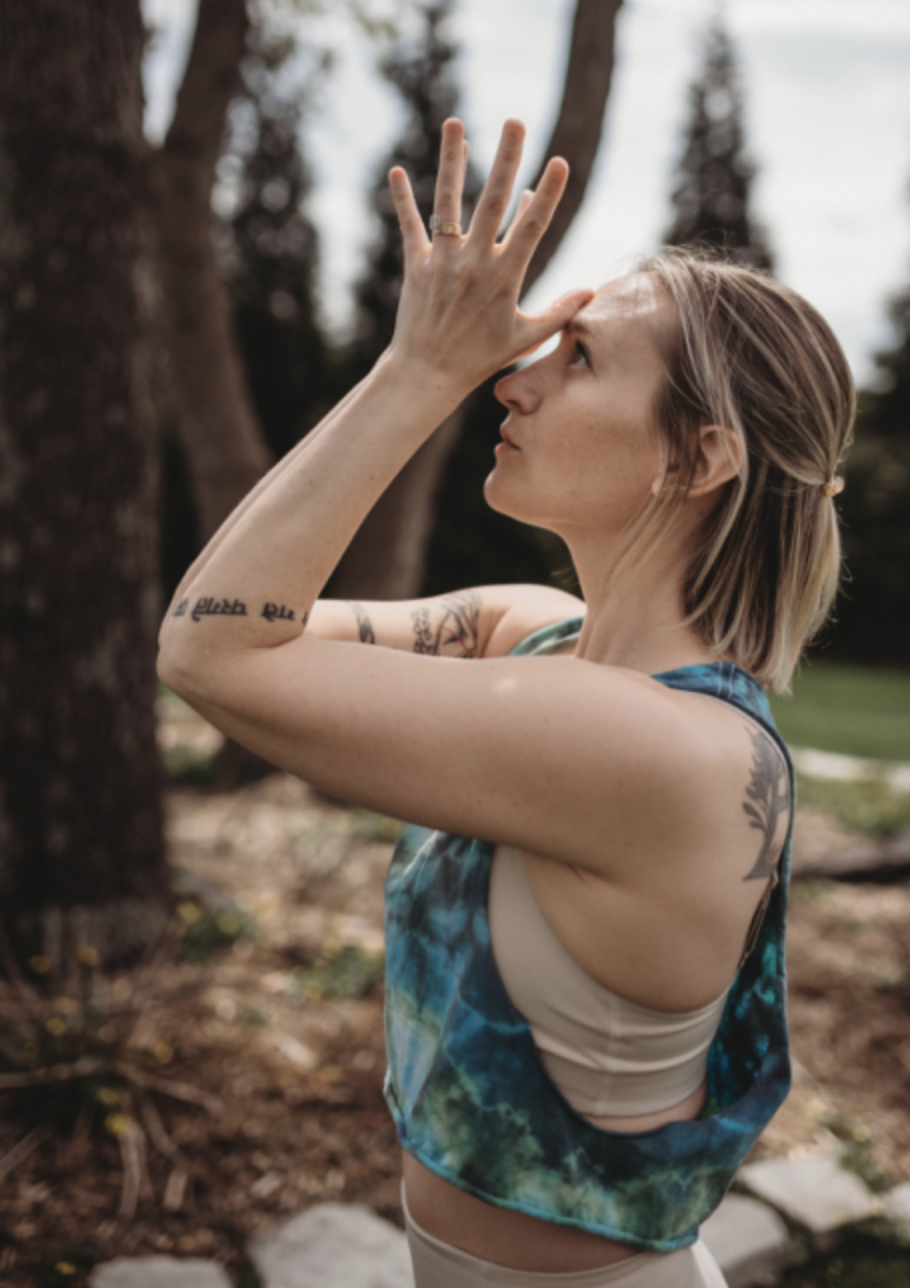 A woman practicing yoga outdoors in a forested area. She is in a prayer pose with her hands pressed together above her forehead, eyes closed, and wearing a colorful sleeveless top and beige sports bra, with tattoos visible on her arms.