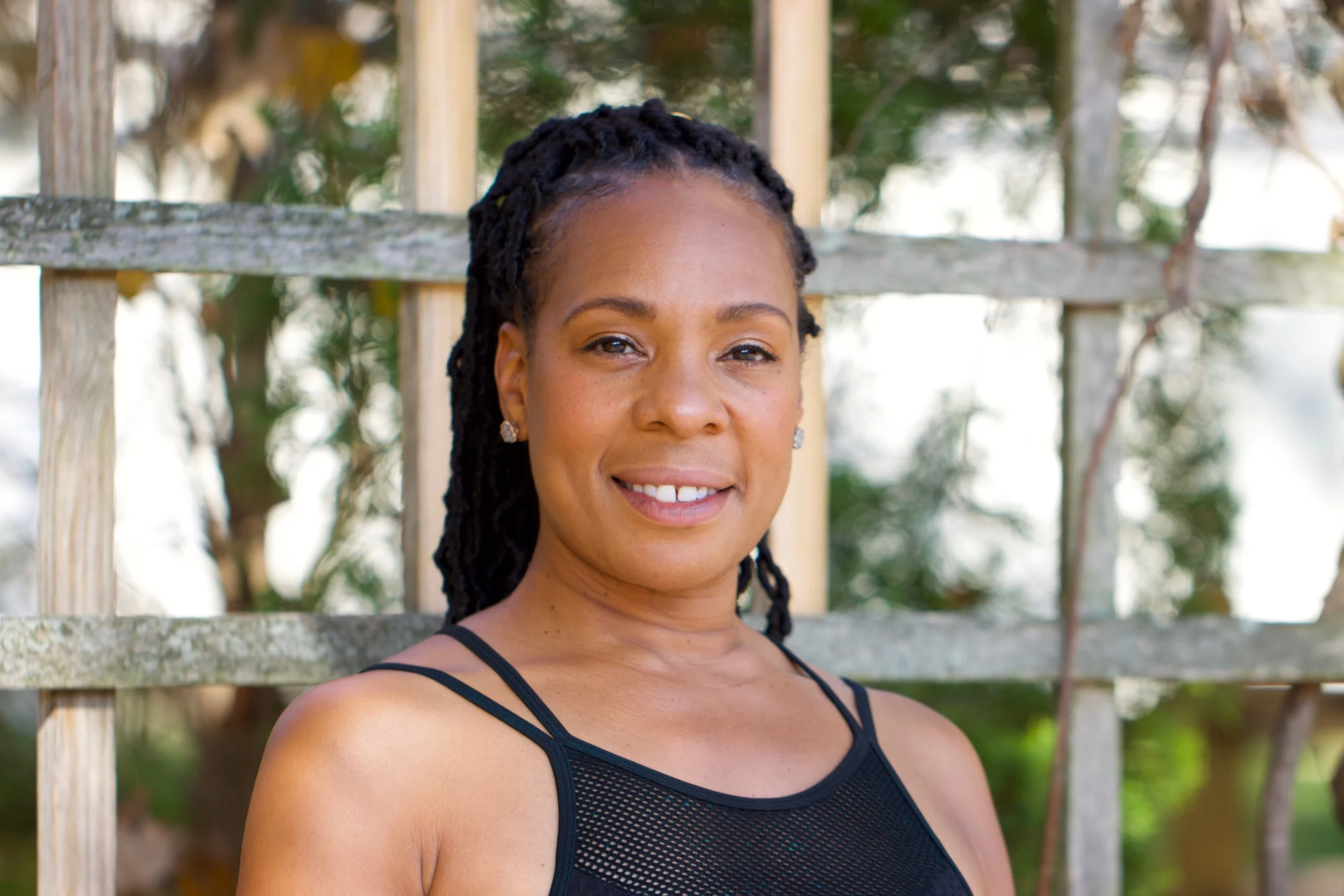 A smiling woman with dreadlocks wearing a black tank top outdoors in front of a wooden fence and greenery.