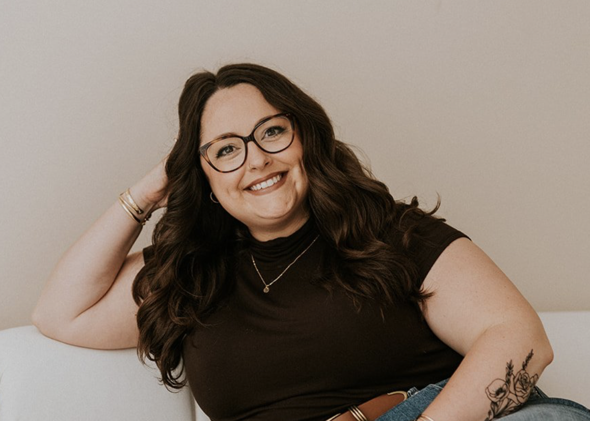 Smiling woman with long curly brown hair, wearing glasses, a black turtleneck top, and jewelry, sitting on a white sofa against a beige wall.