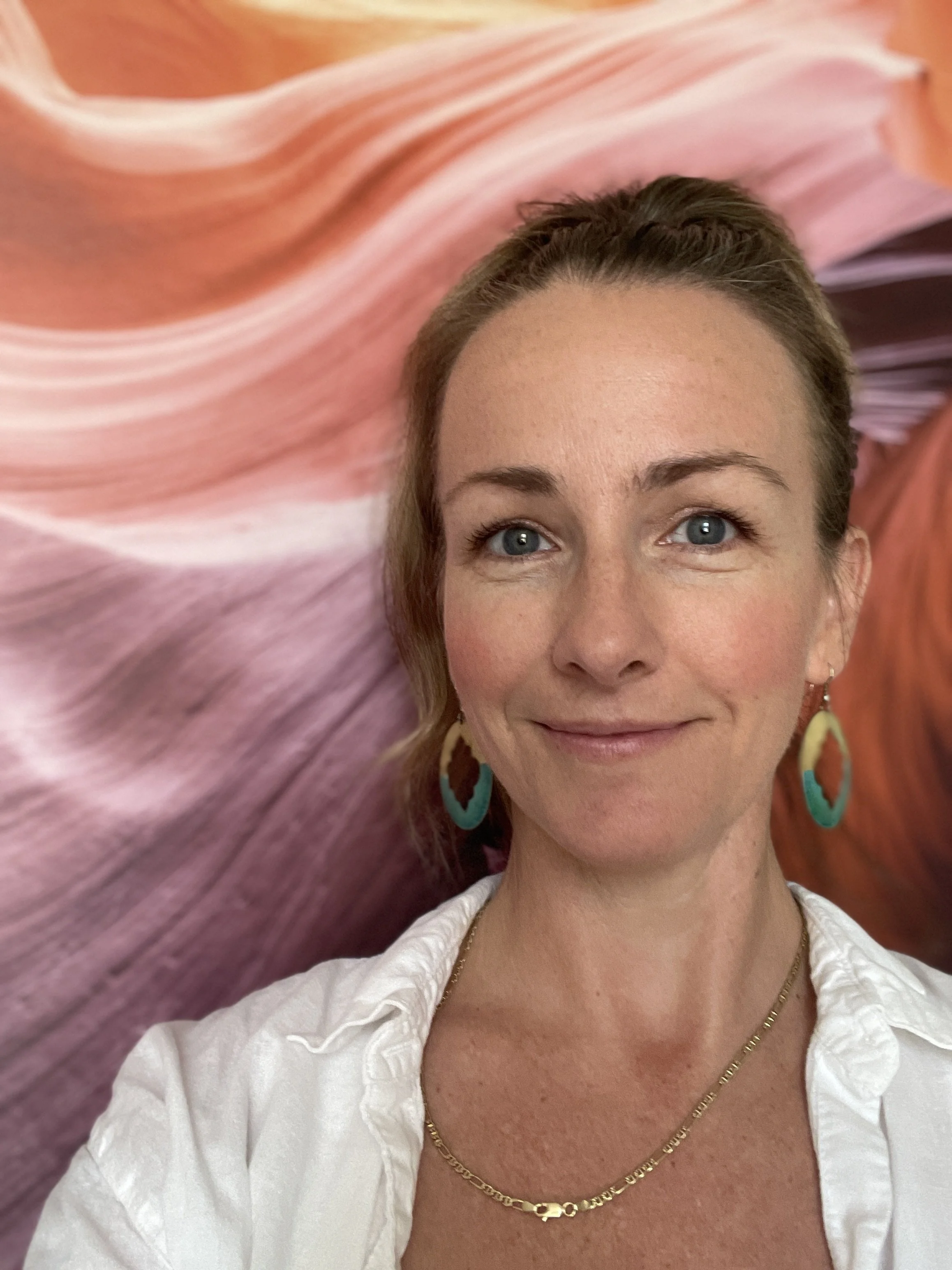 A woman with blue eyes and light brown hair takes a selfie in front of an abstract pink and purple background, wearing colorful earrings and gold jewelry.