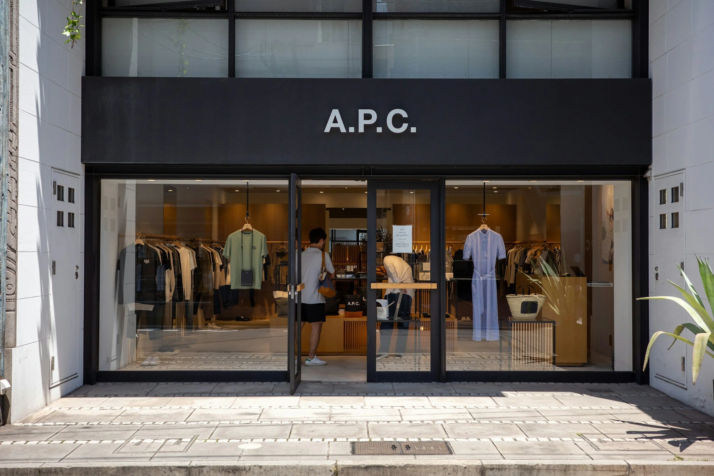 Schaufenster eines Kleidungsgeschäfts namens A.P.C. mit Kleidungsstücken im Inneren und einer Person, die das Geschäft betritt.