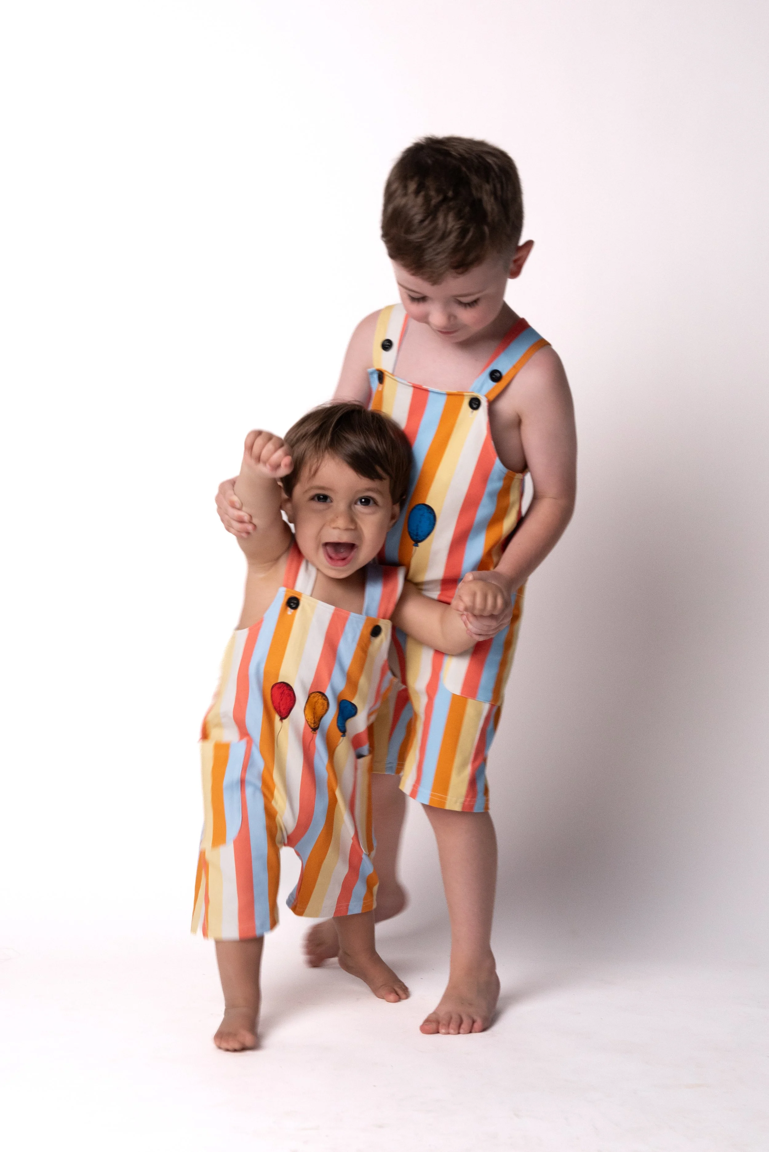 Two young children, a boy and a girl, wearing matching colorful striped overalls with balloon patterns, standing against a plain white background. The girl is smiling and raising her fist, while the boy is looking down at her with a gentle expression.