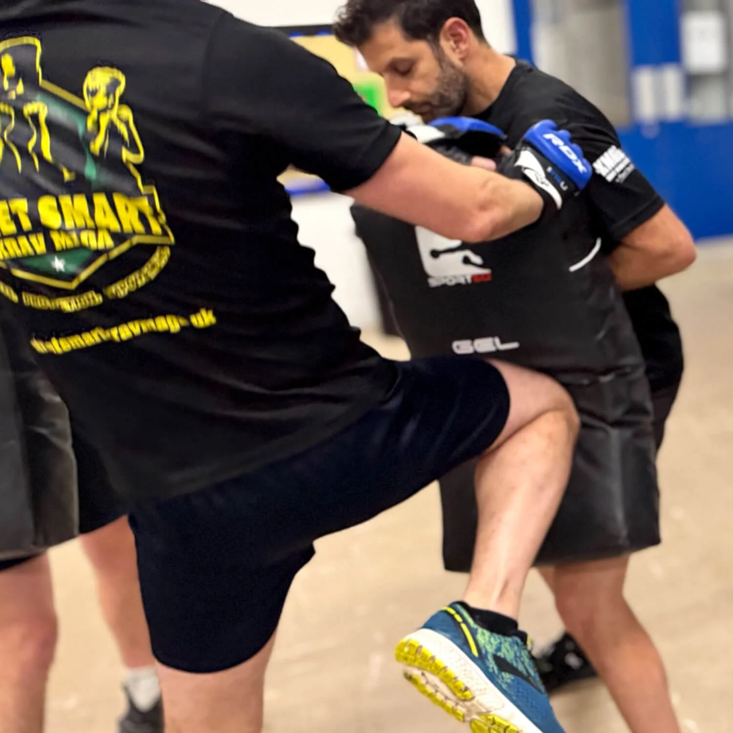 Two men practicing martial arts or boxing, one with a knee raised and the other with gloves, in an indoor gym or training facility.