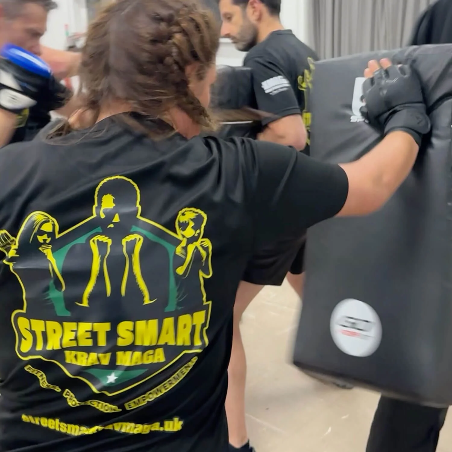 Person wearing a black t-shirt with a yellow and green logo practicing Krav Maga techniques with a punching bag at a gym