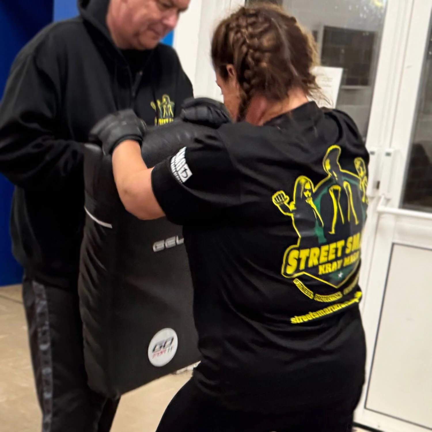 A woman in a black t-shirt and gloves is practicing boxing with a man in black boxing gear, in an indoor training area.