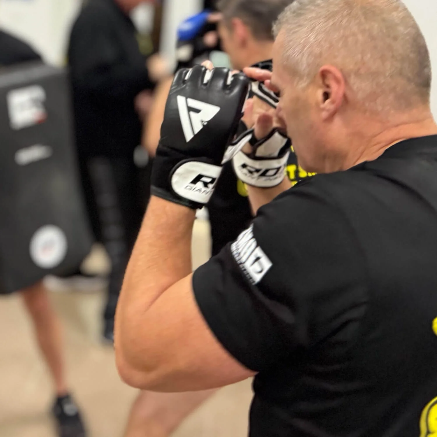 A man wearing a black shirt and gloves preparing for a mixed martial arts fight in a gym, with other fighters in the background.