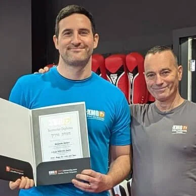 Two men standing side by side, smiling. The man on the left is holding an open certificate or award. Both are wearing gray T-shirts with a logo and text. Red fitness equipment is visible in the background.