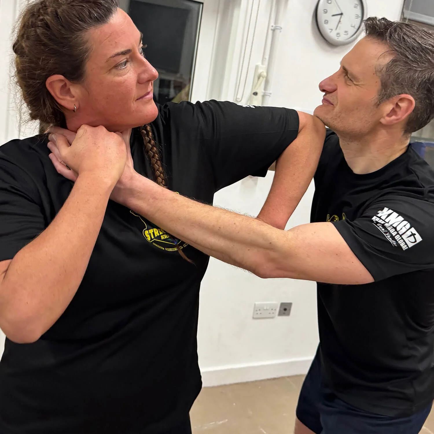 A woman and a man are engaged in a playful wrestling match indoors, with the woman grabbing the man's chest with one arm and his neck with the other, both wearing black shirts with logos.