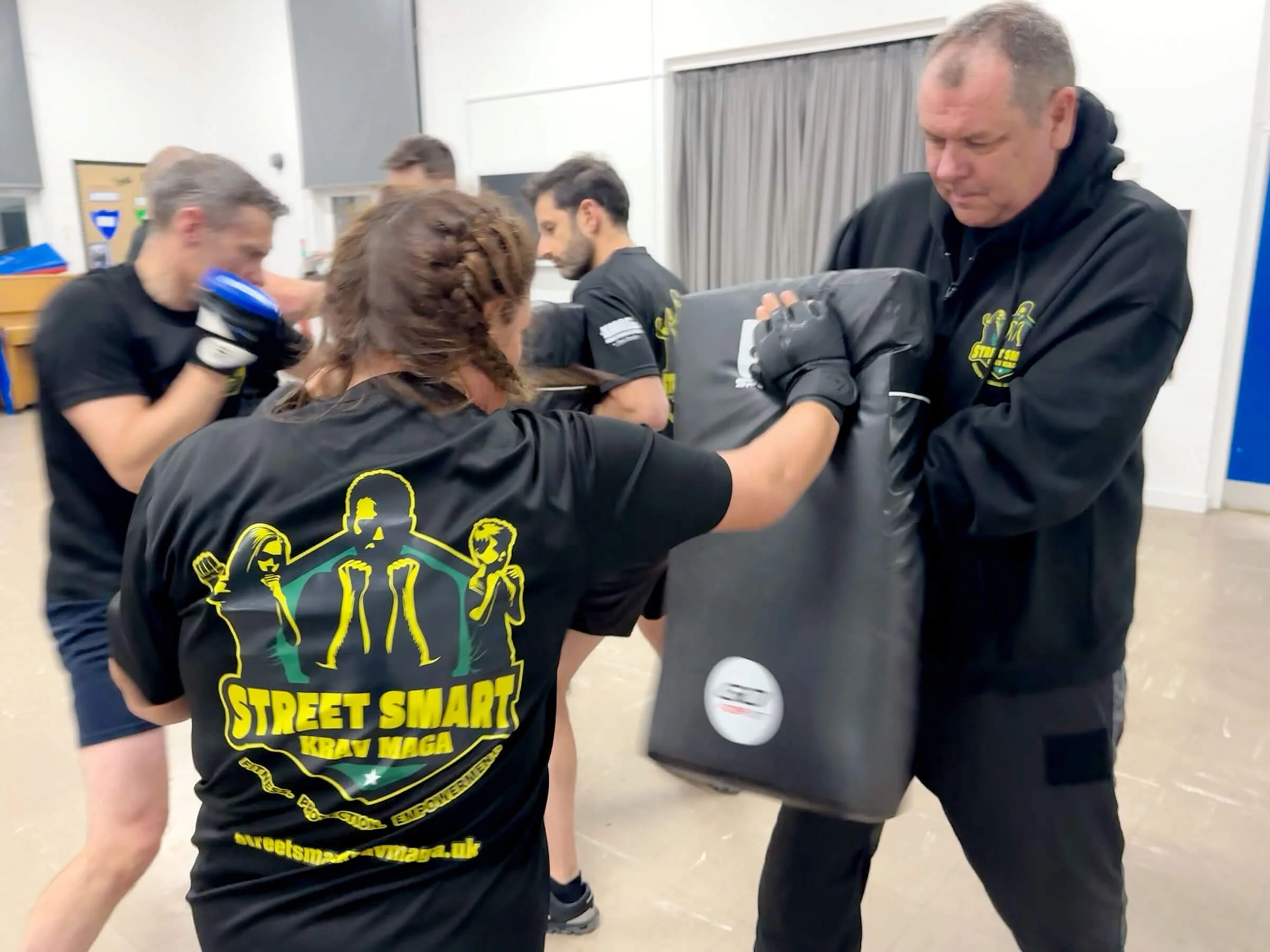 Group of people practicing Krav Maga, wearing black T-shirts with the logo 'Street Smart Krav Maga', in a dojo, with one person practicing punches on a padded shield held by another person.