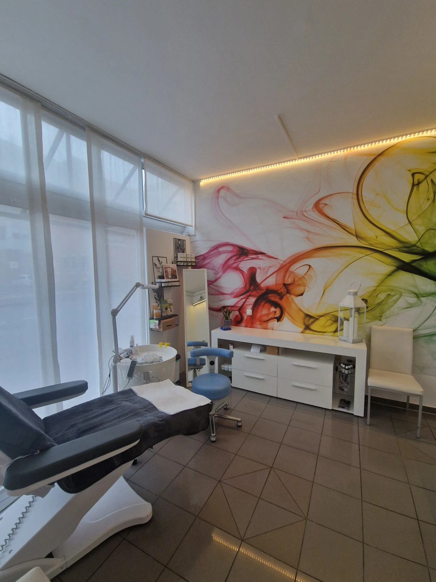 Ein Behandlungsraum in einer modernen dermatologischen Praxis mit einem Behandlungstisch, Stuhl, medizinischer Ausrüstung und einer Wand mit buntem, abstraktem Wandbild.