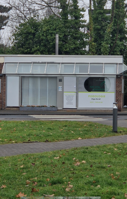 Außenansicht einer Podologie-Praxis in einem kleinen Gebäude mit großen Fenstern, einem grauen Vorhang und einem weißen Schild mit grüner Schrift. Im Hintergrund sind Bäume, im Vordergrund ein grüner Rasen mit einem Gehweg und einem Strommast.