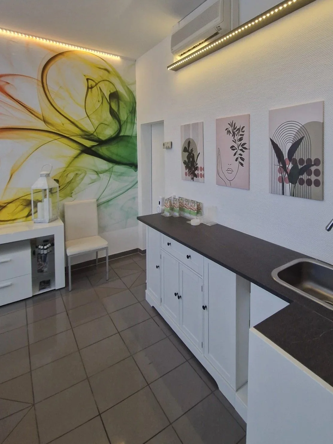 Ein Raum mit weißer Wand, auf der drei moderne Kunstwerke mit abstrakten Frauengesichtern hängen. Die linke Wand hat eine große, bunte Wandmalerei mit grünen, gelben und braunen Farbverläufen. Es gibt eine weiße Sitzbank mit einem weißen Lampenständer daneben, sowie einen weißen Küchenschrank mit schwarzer Arbeitsplatte und Spüle.