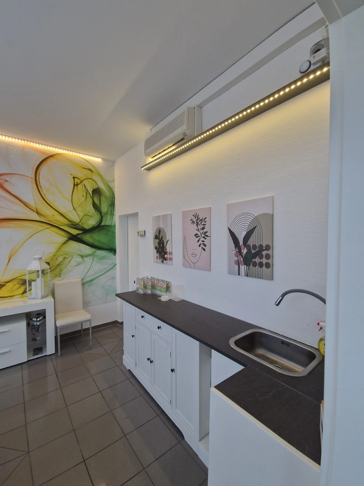 Innenraum mit weißen Wänden, dekorativer Wandkunst, Küchenbereich mit dunklem Arbeitsplatten, Spüle, Kaffeemaschine, Stühlen und einer bunten Wandgestaltung in Akzentfarben.