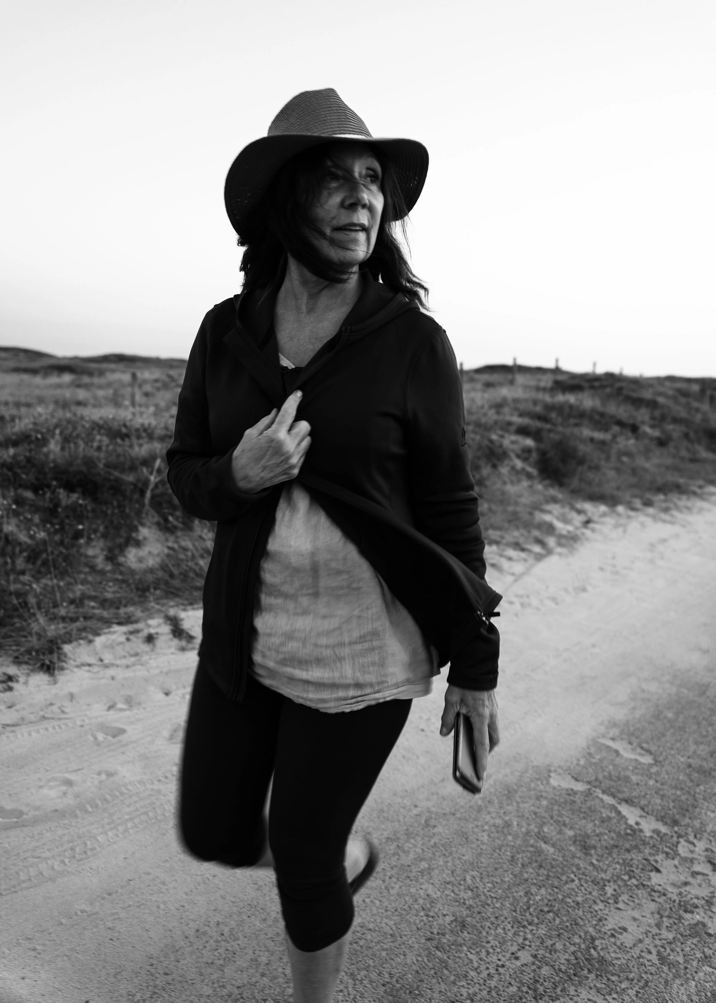 Une femme portant un chapeau et une veste noire marche sur un chemin de sable dans un paysage naturel, en noir et blanc.